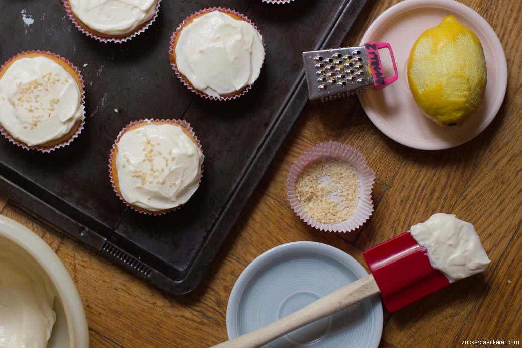 cupcakes mit frosting, daneben eine zitrone mit zitronenreibe, ein silikonspatel mit frosting und ein kleines schüsselchen mit sesamsamen, vogelperspektive