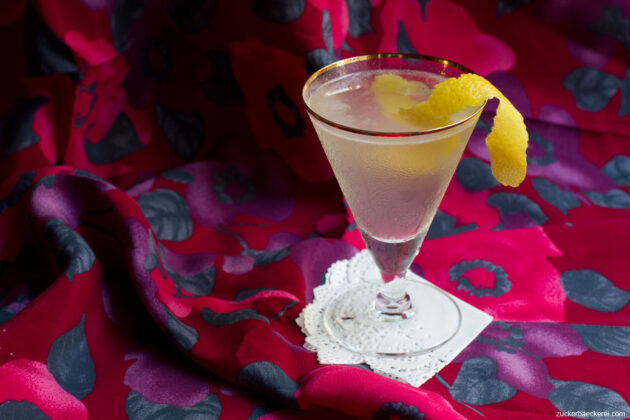 martiniglas mit leicht trübem hellen cocktail und einer zitronenzeste, im hintergrund eine grob geblümte tischdecke