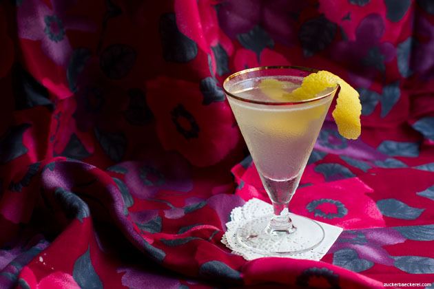 martiniglas mit leicht trübem hellen cocktail und einer zitronenzeste, im hintergrund eine grob geblümte tischdecke